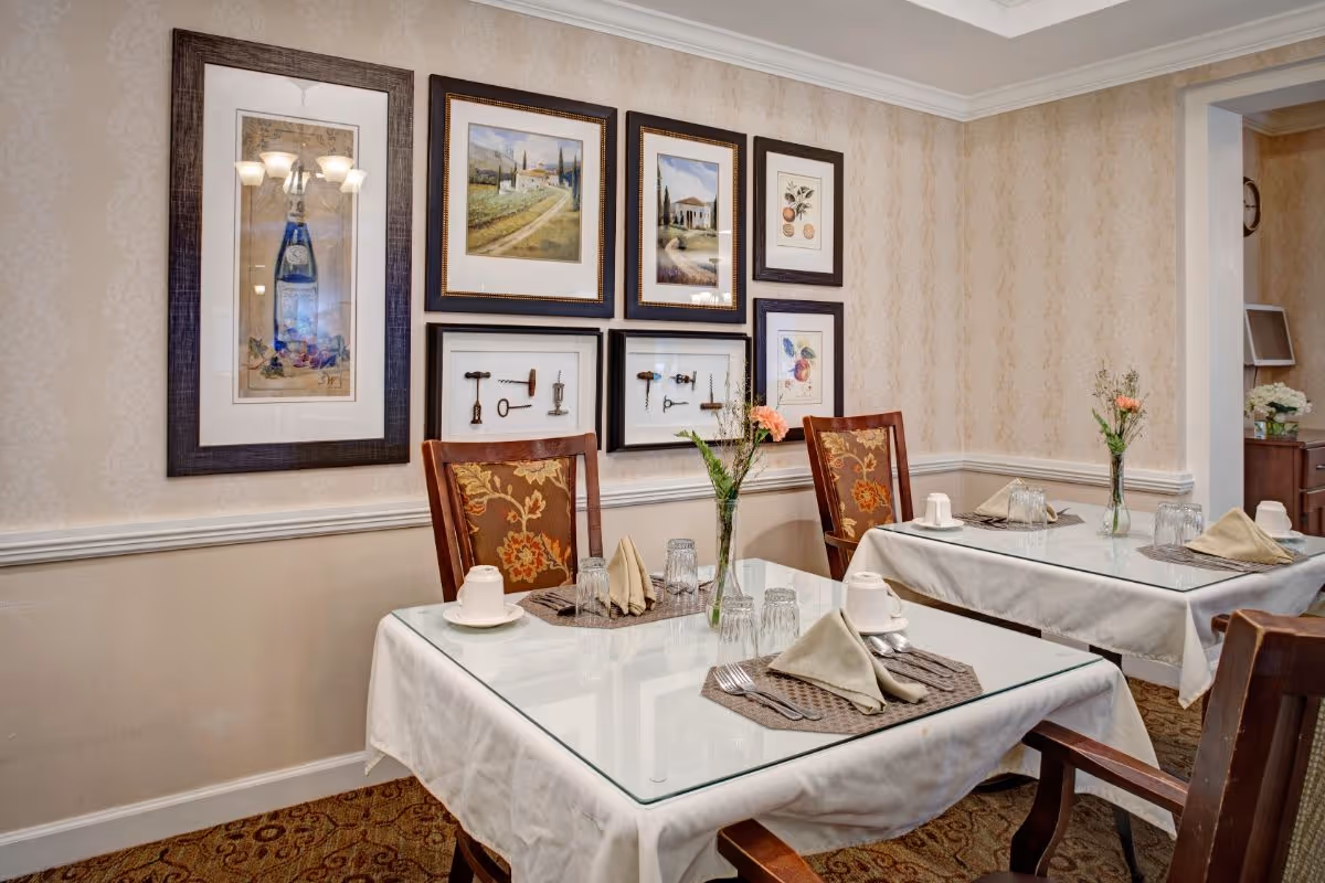 A cozy dining area with two tables covered in white tablecloths and glass tops, each set with beige napkins, cups, glasses, and silverware. The chairs have wooden frames with floral patterned cushions. The walls are decorated with framed artwork including paintings and vintage keys. A vase with flowers is placed on each table.