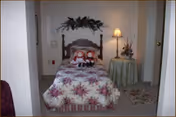 Small single bedroom with a floral bedspread and two dolls on the bed, a wooden headboard, and a round side table with a lamp.