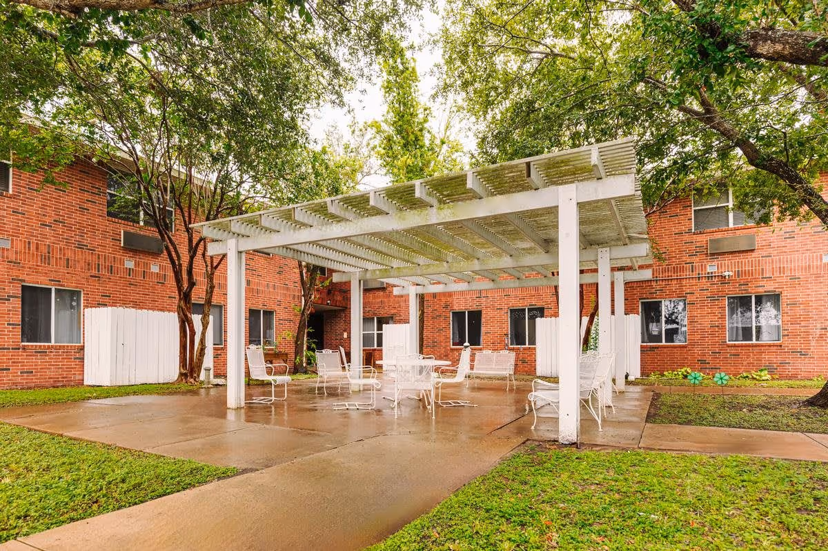 Outdoor courtyard with a white pergola and metal patio chairs in front of a red-brick senior living building.