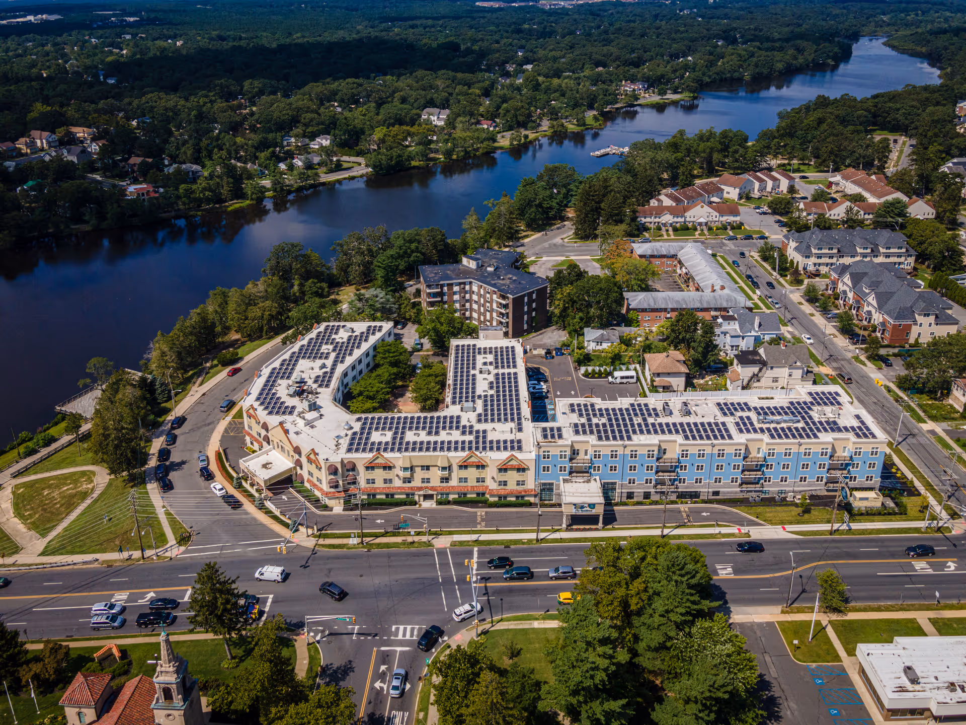 Aerial view of the Lakewood Courtyard senior living complex with solar-panel-covered rooftops beside a river and surrounding neighborhood.
