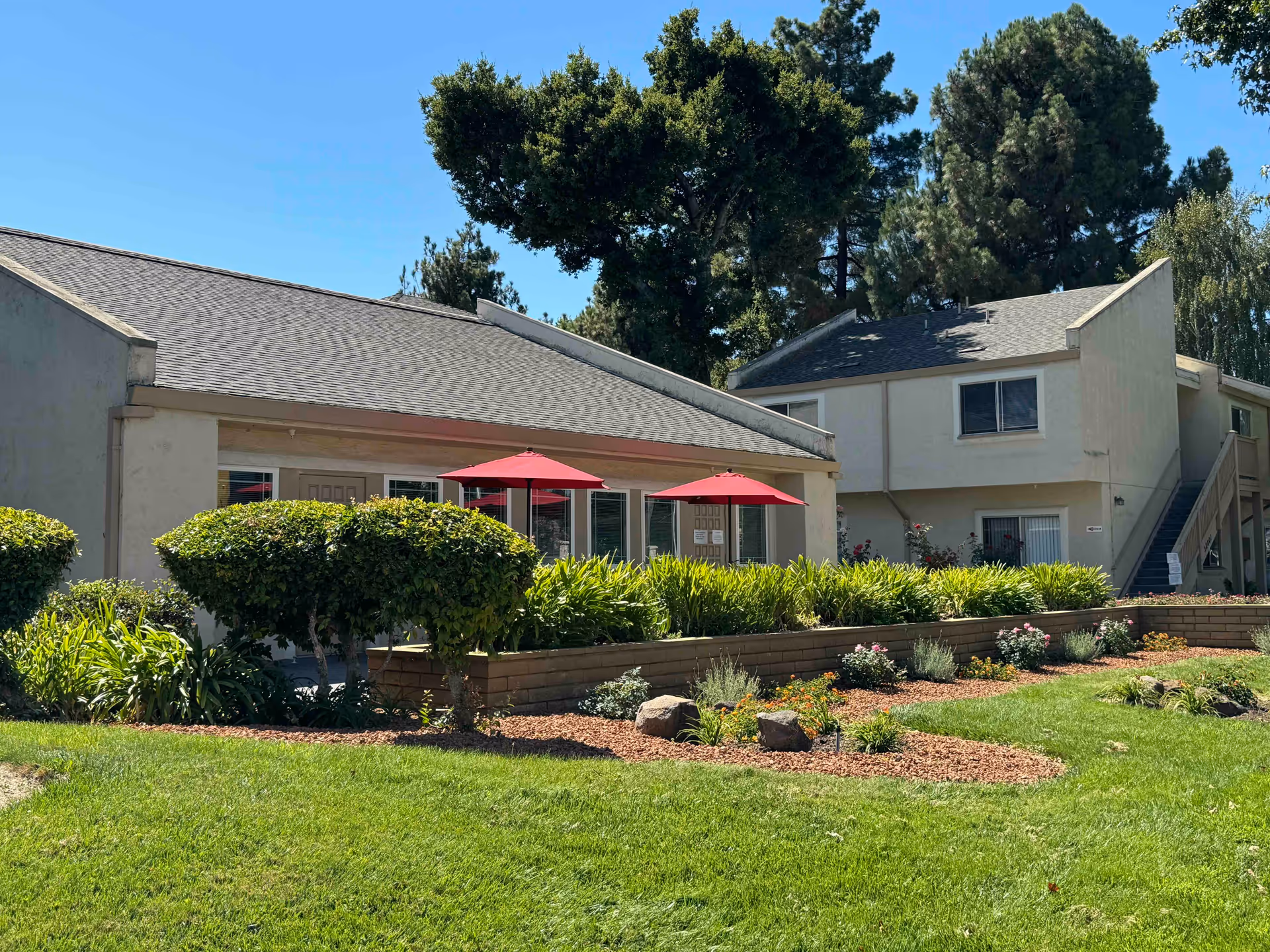 Outdoor view of a senior living facility named Fremont Village featuring well-maintained green lawns, trimmed bushes, flower beds with mulch, and two red patio umbrellas in front of a single-story building. There is also a two-story building with an external staircase in the background, surrounded by tall trees under a clear blue sky.