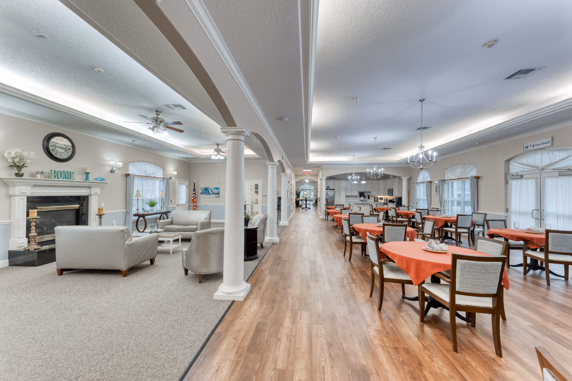 A spacious and well-lit common area in a senior living facility featuring a living room section with beige sofas, a fireplace, and decorative items on the mantel. Adjacent to the living room is a dining area with multiple tables covered with orange tablecloths and chairs arranged neatly. The room has wooden flooring, chandeliers, ceiling fans, and large windows with curtains allowing natural light to enter.