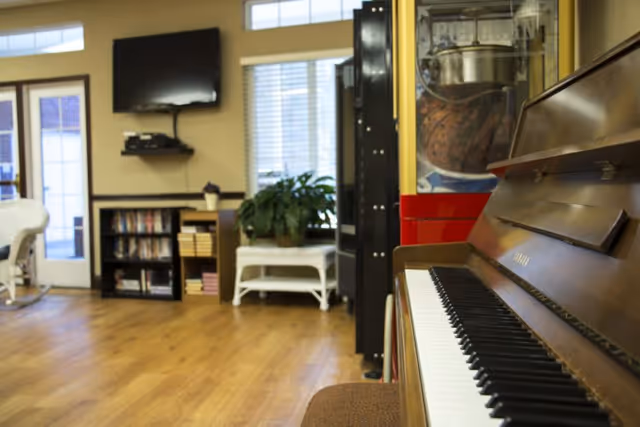 Interior of a senior living facility common area with a wooden piano in the foreground, a popcorn machine, a TV mounted on the wall, bookshelves, a white wicker bench with a plant, and large windows letting in natural light.