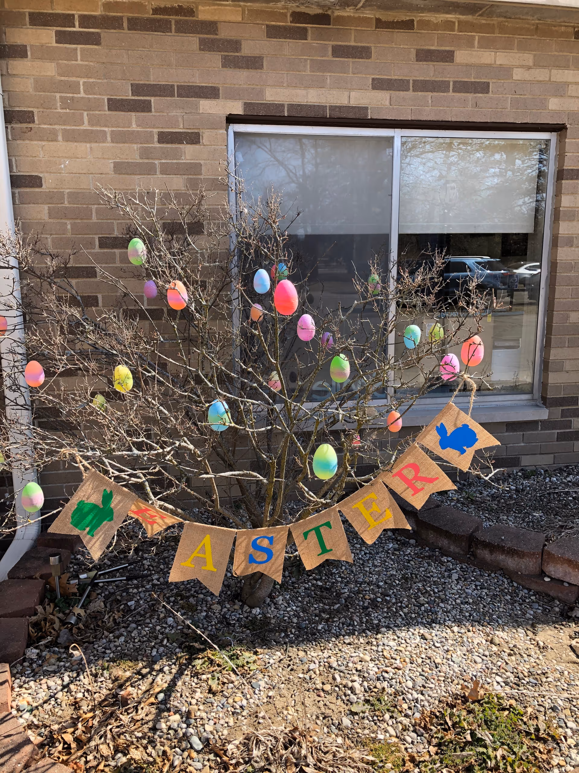 A leafless shrub outside a brick building is decorated with colorful hanging Easter eggs and a burlap banner spelling EASTER.