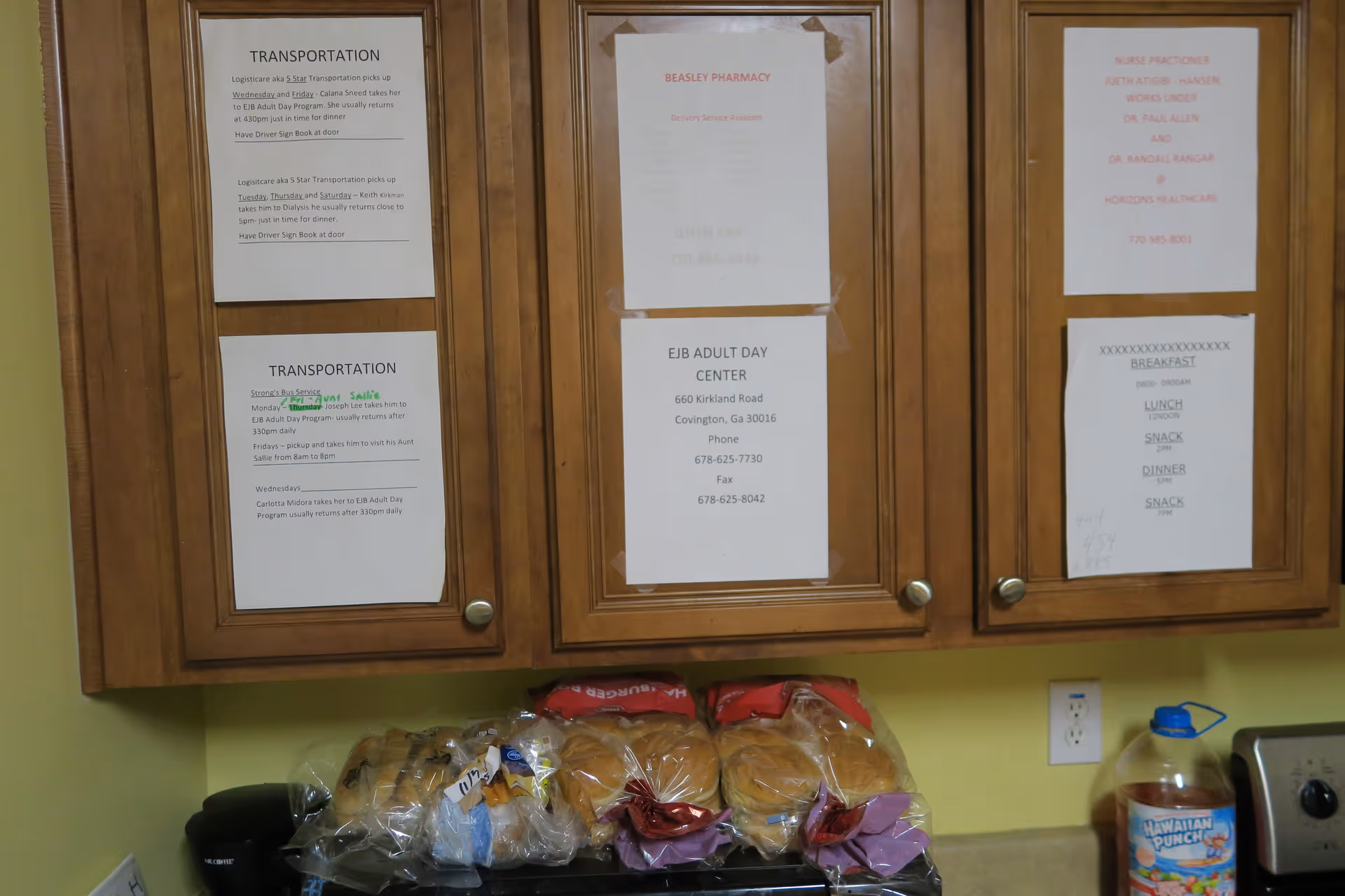 Wooden kitchen cabinets with multiple printed notices taped on the doors. Below the cabinets, there are several packages of bread and buns on a countertop, along with a large bottle of Hawaiian Punch and a toaster oven. The wall is painted light green.