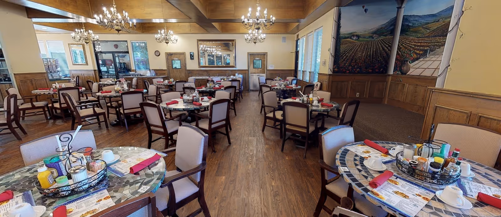 A spacious dining room with multiple round tables set with menus, red napkins, cups, and condiment baskets. The room features wooden floors, beige walls with wood paneling, large windows with blinds, chandeliers hanging from a wooden beam ceiling, and a large mural painting on one wall.