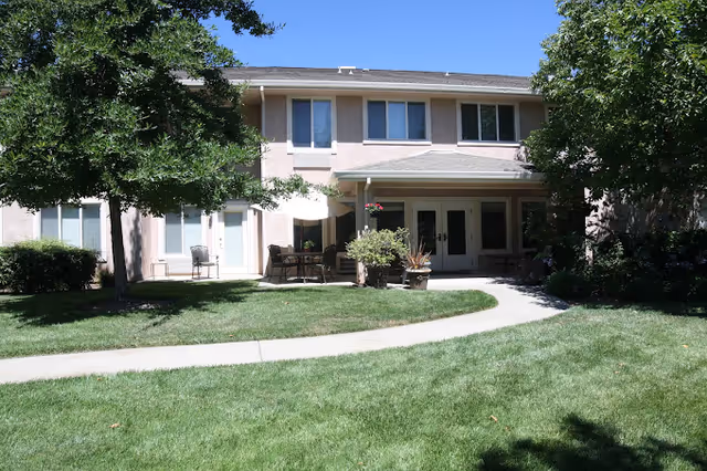 Two-story beige building with a covered patio, outdoor seating, potted plants, trees, and a curved walkway across a green lawn.