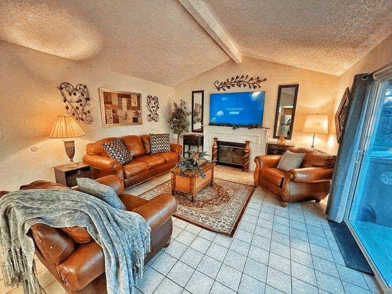 A cozy living room with three brown leather seating pieces including a sofa and two armchairs. The room features a tiled floor with a patterned area rug, a coffee table with a decorative centerpiece, a fireplace with a mounted flat-screen TV above it, two tall mirrors on either side of the fireplace, and warm lighting from two table lamps. There is a sliding glass door with curtains on the right side leading to an outdoor area.