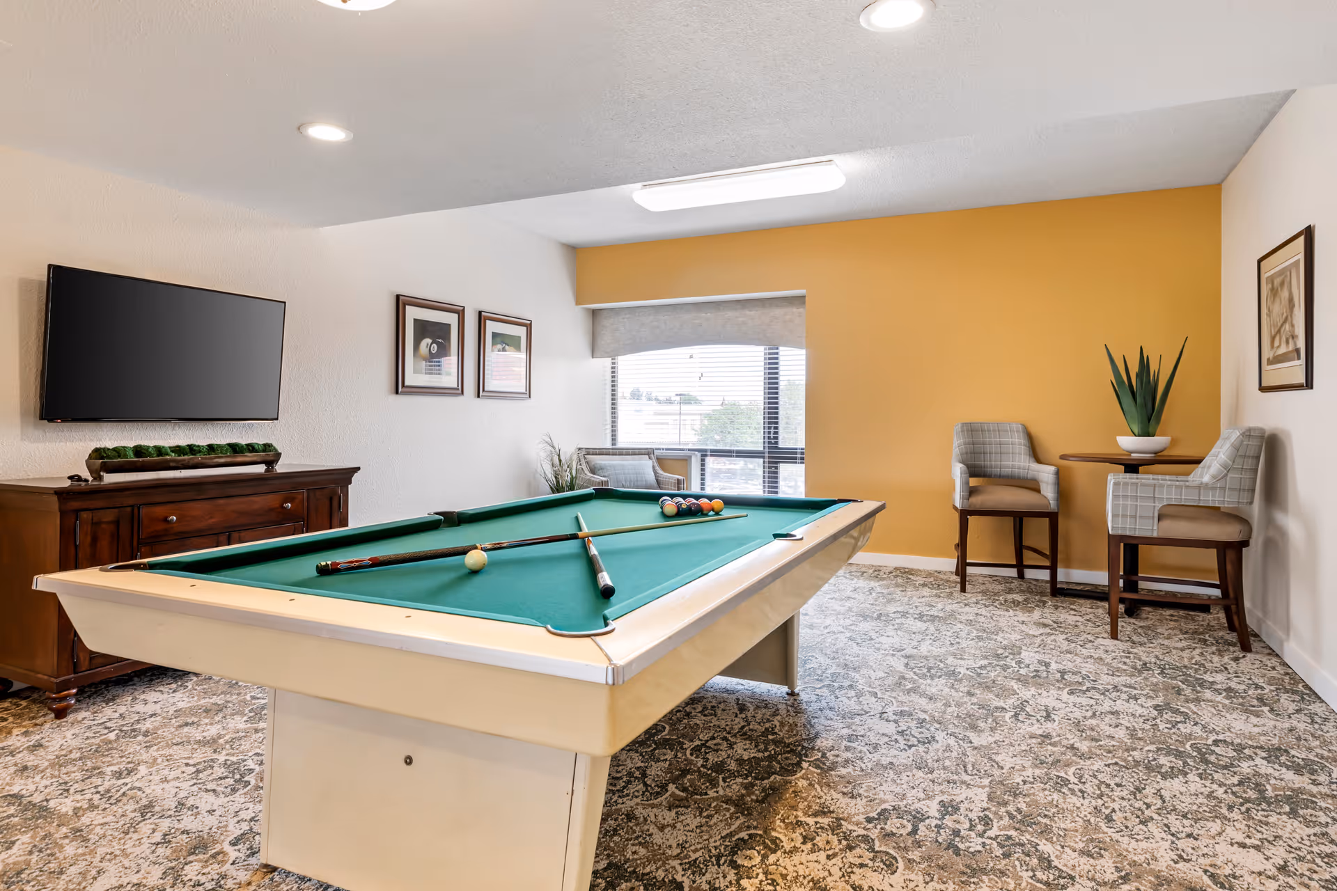 A recreational room with a pool table in the center, two pool cues and balls arranged on the table. The room has a patterned carpet, a flat-screen TV mounted on the wall above a wooden cabinet, two framed pictures on the wall, a window with blinds, and a small seating area with two upholstered chairs and a round table with a plant on it.