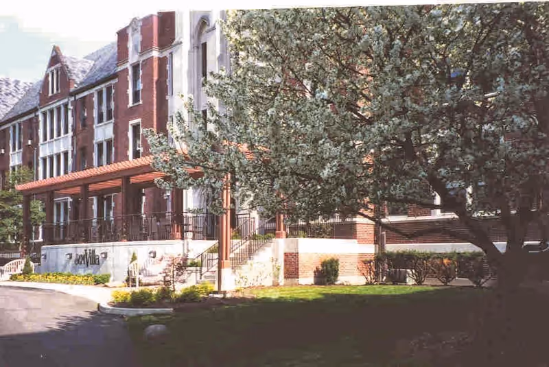 Front entrance of a multi-story brick senior living building with a covered porch and large tree in the foreground.
