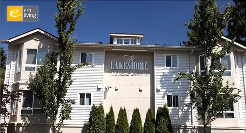 Exterior view of The Lakeshore Retirement Residence building with white siding, multiple windows, and trees in front under a clear blue sky.