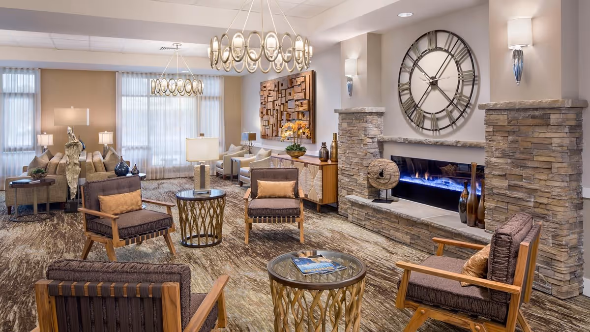 A spacious and well-lit senior living facility lounge area featuring comfortable wooden armchairs with cushions, round glass-top tables, a stone fireplace with a modern electric fire, and a large decorative clock above the fireplace. The room has large windows with sheer curtains, contemporary chandeliers, and decorative wall art, creating a warm and inviting atmosphere.