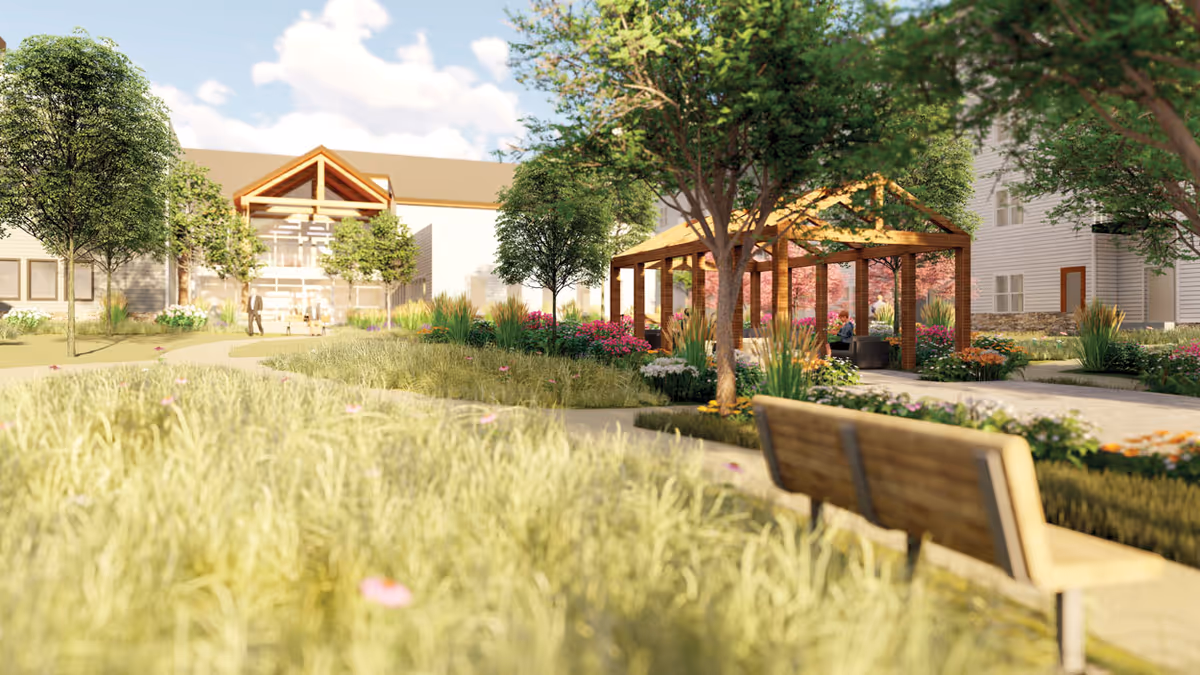 Outdoor garden area at Benedictine Living Community- Shakopee Windermere Way featuring a wooden bench in the foreground, a paved walkway, a wooden pergola with seating underneath, surrounded by trees, flowering plants, and grass, with the building visible in the background under a partly cloudy sky.