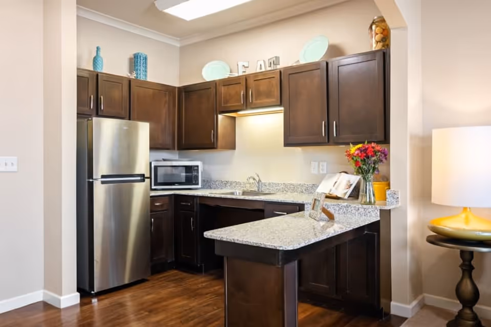 Small kitchen with dark wood cabinets, stainless steel refrigerator, microwave, granite countertops and a vase of flowers on the breakfast bar.
