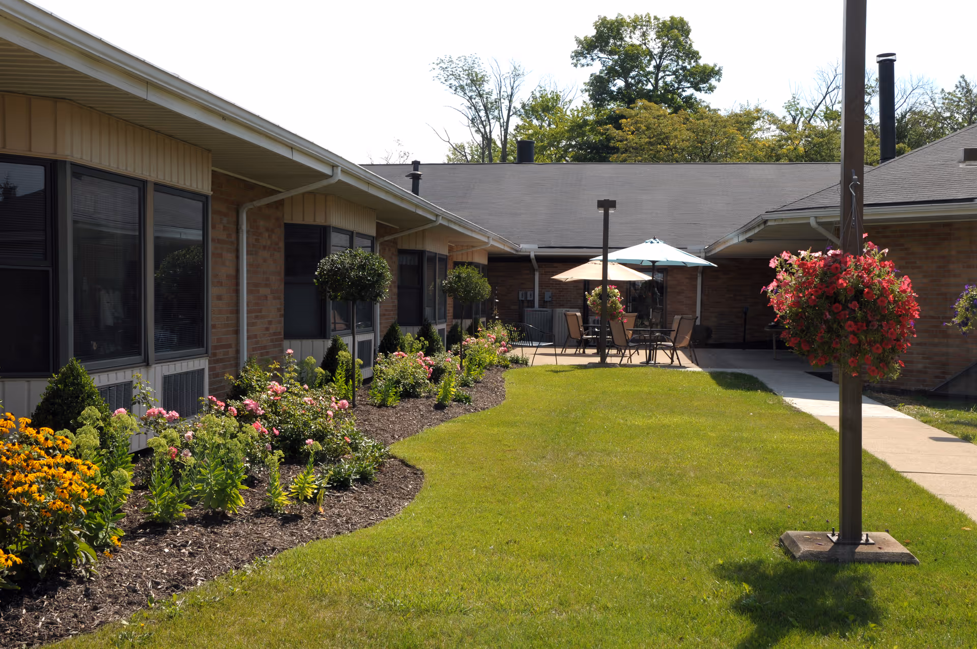 A sunny courtyard between single-story brick buildings with manicured flower beds, a lawn, patio tables with umbrellas, and hanging flower baskets.