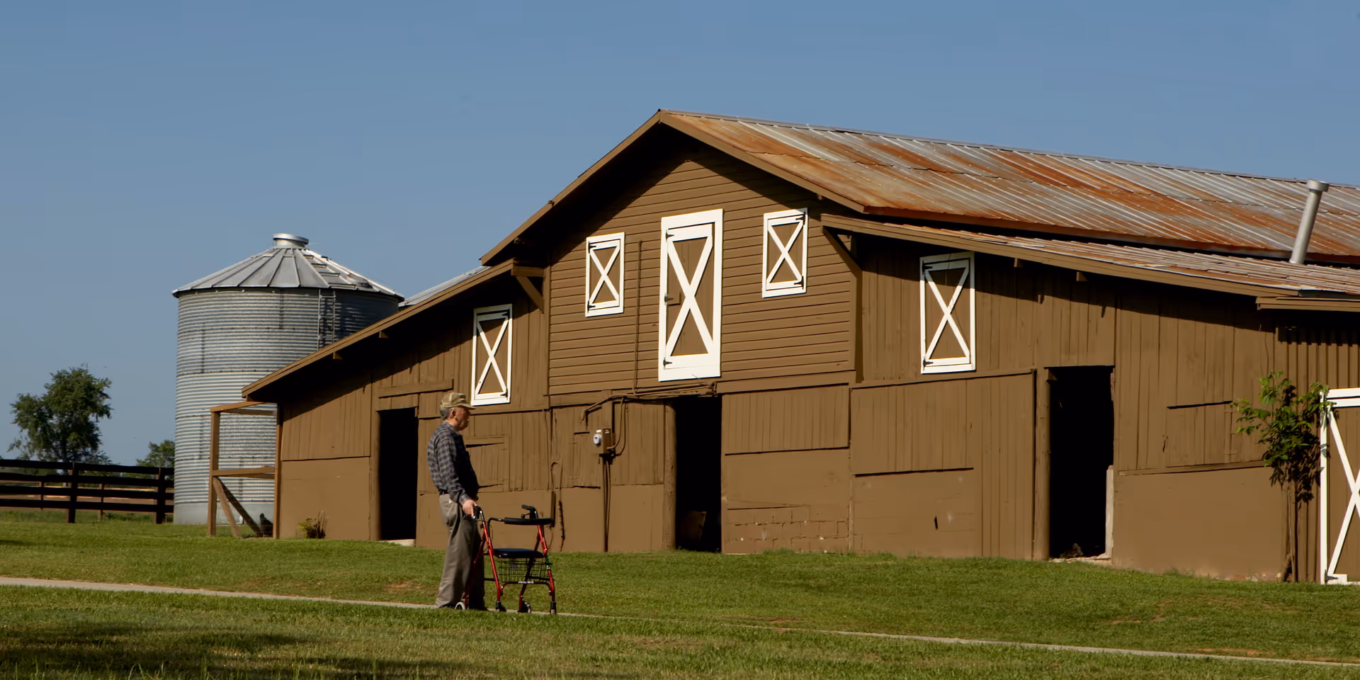 An elderly man using a walker is standing on a grassy area near a large brown barn with white-trimmed windows and doors. A metal silo and a wooden fence are visible in the background under a clear blue sky.