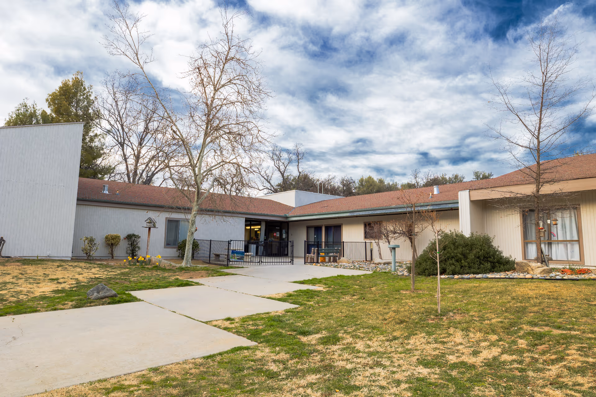 Front exterior of a single-story senior care building with a paved walkway, lawn, bare trees, and a cloudy sky overhead.