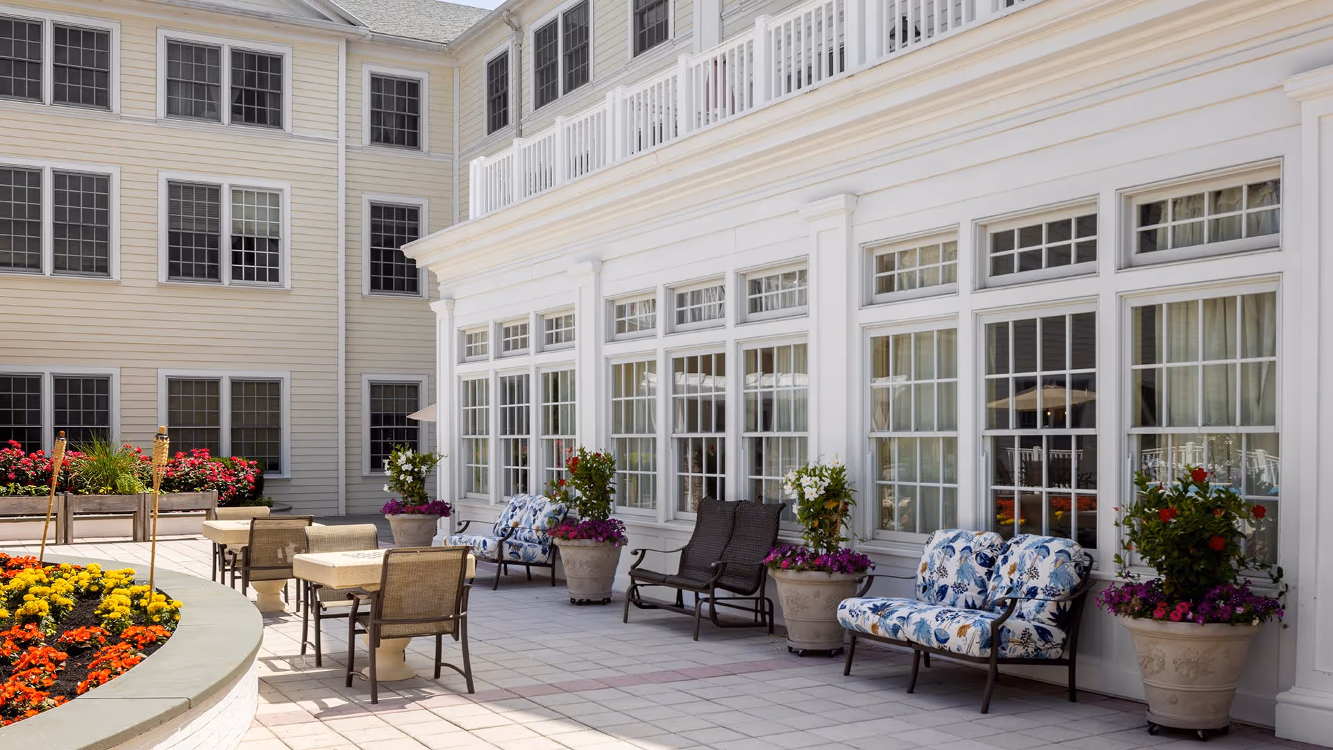 Outdoor patio area at a senior living facility with several tables and chairs arranged on a tiled floor. The patio is adjacent to a building with large white-framed windows and white siding. There are multiple large flower pots with colorful flowers and greenery placed along the building wall. A curved flower bed with vibrant yellow, red, and orange flowers is visible in the foreground.