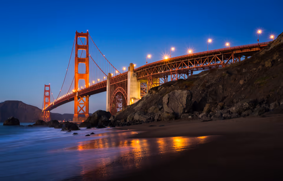 The Golden Gate Bridge lit at dusk with its lights reflecting on the wet sand and ocean in the foreground.