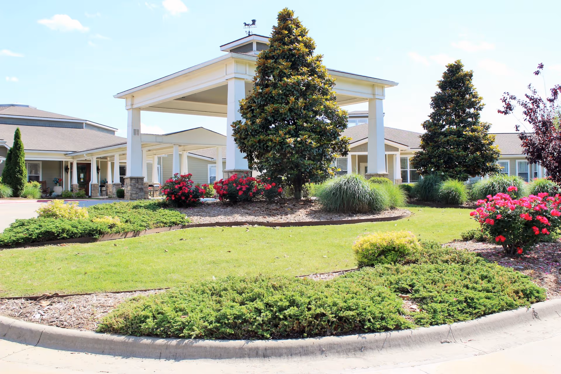 Front exterior of a senior living facility with a covered porte-cochere, manicured lawn, flowering bushes and trees.
