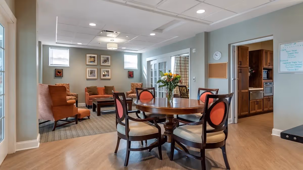 A cozy senior living facility common area with a round wooden table and four chairs with red cushions in the foreground. Behind the table is a seating area with armchairs and a coffee table, framed pictures on the wall, and two small windows letting in natural light. To the right, there is an open doorway leading to a kitchen with wooden cabinets and appliances. The room has light-colored walls and wood flooring.