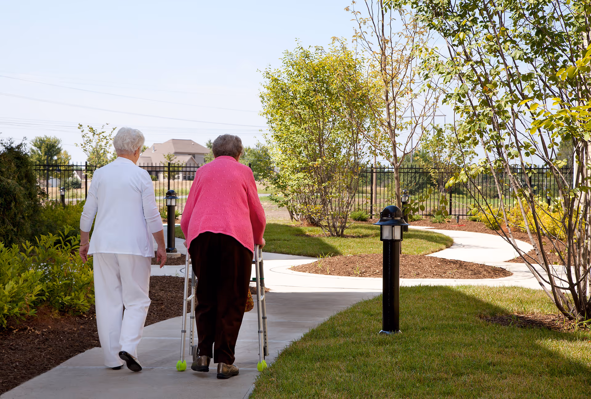 Two older adults walking along a curved paved path in a landscaped outdoor area, one using a walker.