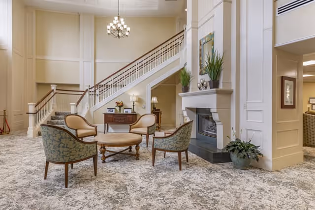 Bright lobby with upholstered chairs around a round table, a fireplace, and a grand staircase under a chandelier.