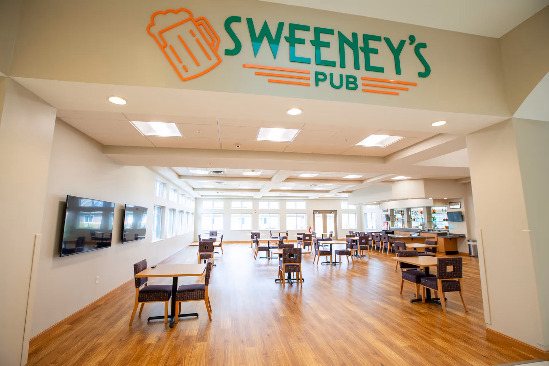 Interior view of Sweeney's Pub, a spacious and well-lit room with wooden flooring, multiple tables and chairs arranged neatly, large windows letting in natural light, a bar area on the right side, and two flat-screen TVs mounted on the left wall.