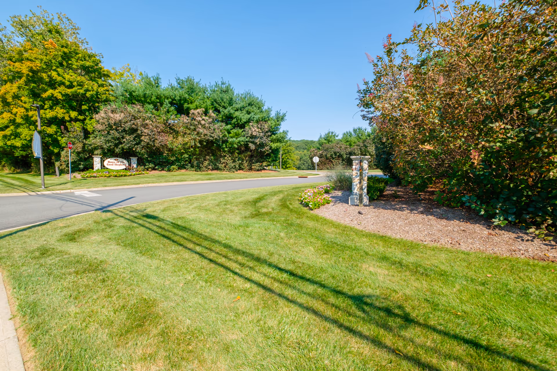 Landscaped entrance to a senior living community with a curved road, manicured lawn, trees, and a small stone entrance sign.