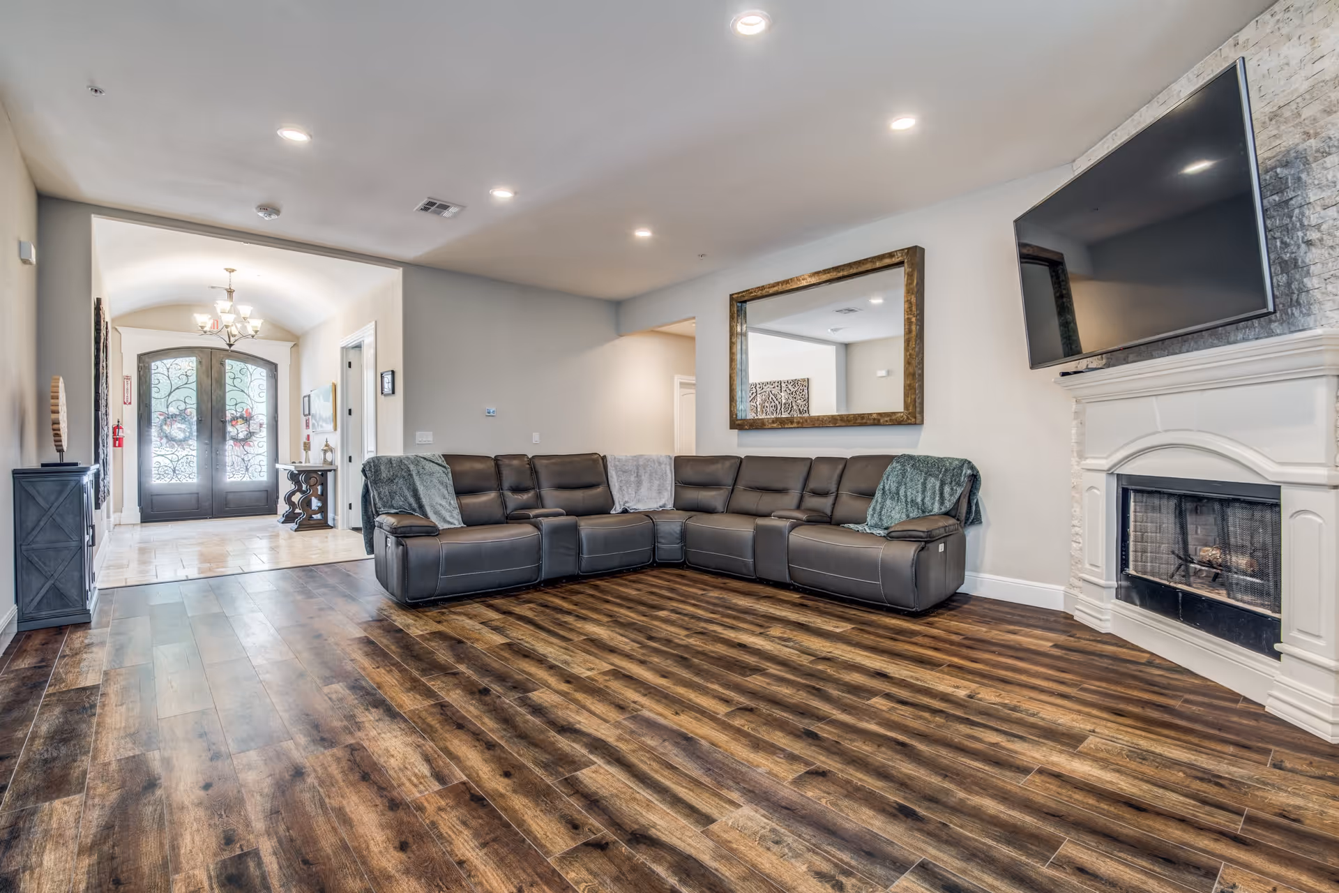A spacious living room with a large dark brown leather sectional sofa adorned with three blankets. The room features wood flooring, a white fireplace with a mounted flat-screen TV above it, and a large rectangular mirror on the wall. In the background, there is an entryway with double doors featuring decorative glass and wrought iron designs, a chandelier, and a small table with decor items.