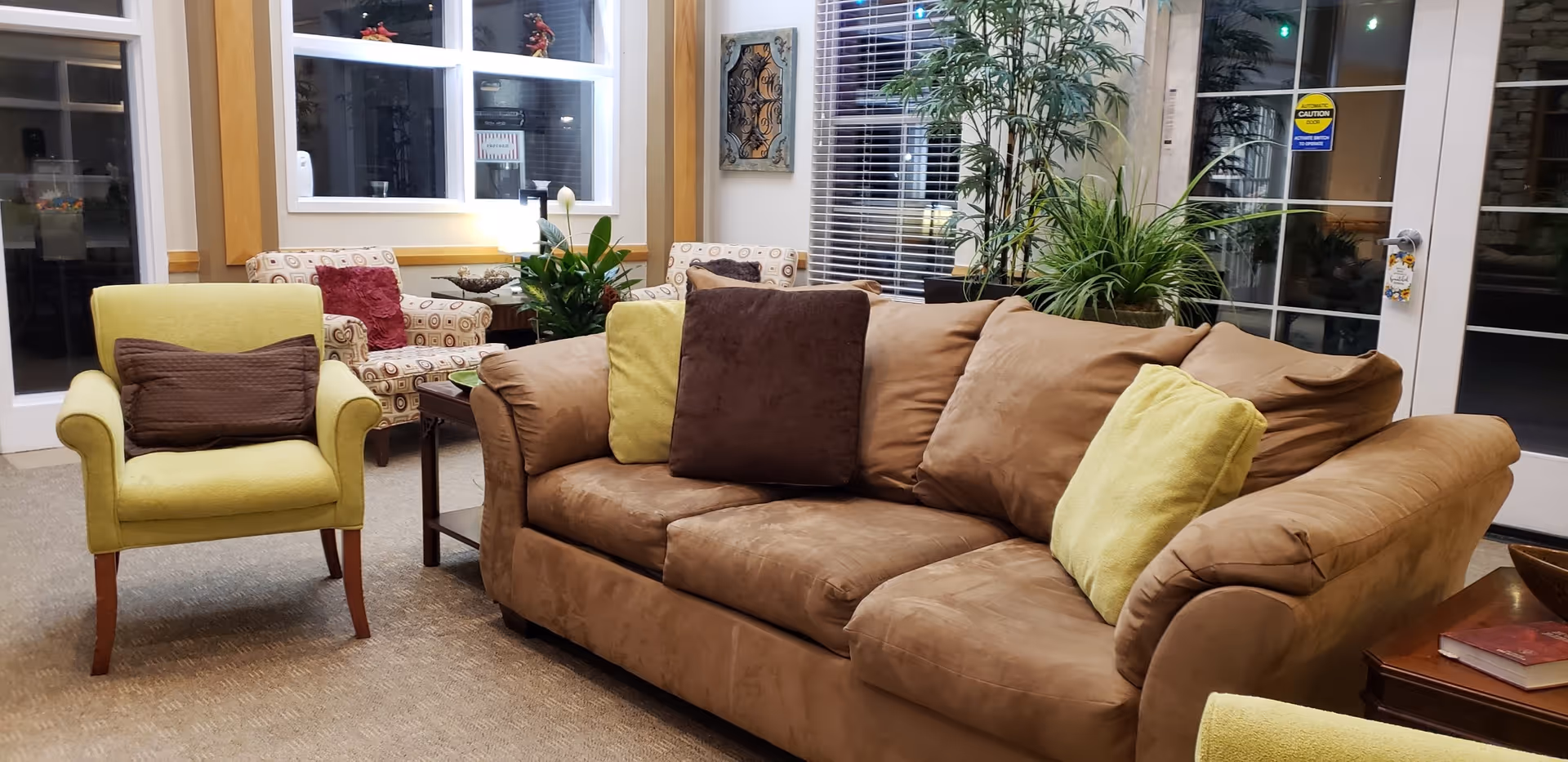 A cozy living room area with a brown sofa adorned with green and brown cushions, a green armchair with a brown pillow, two patterned armchairs, a wooden side table with a plant, and large windows with blinds. There are decorative plants and wall art visible, creating a warm and inviting atmosphere.