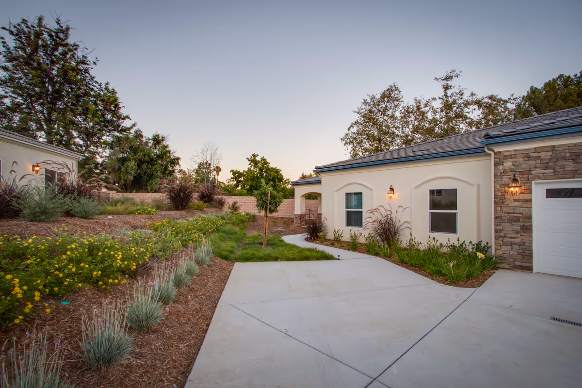 Exterior view of a senior living facility showing a paved driveway leading to a building with cream-colored walls, stone accents, and outdoor wall lanterns. The landscaped area beside the driveway features various plants, shrubs, and small trees under a clear sky at dusk.