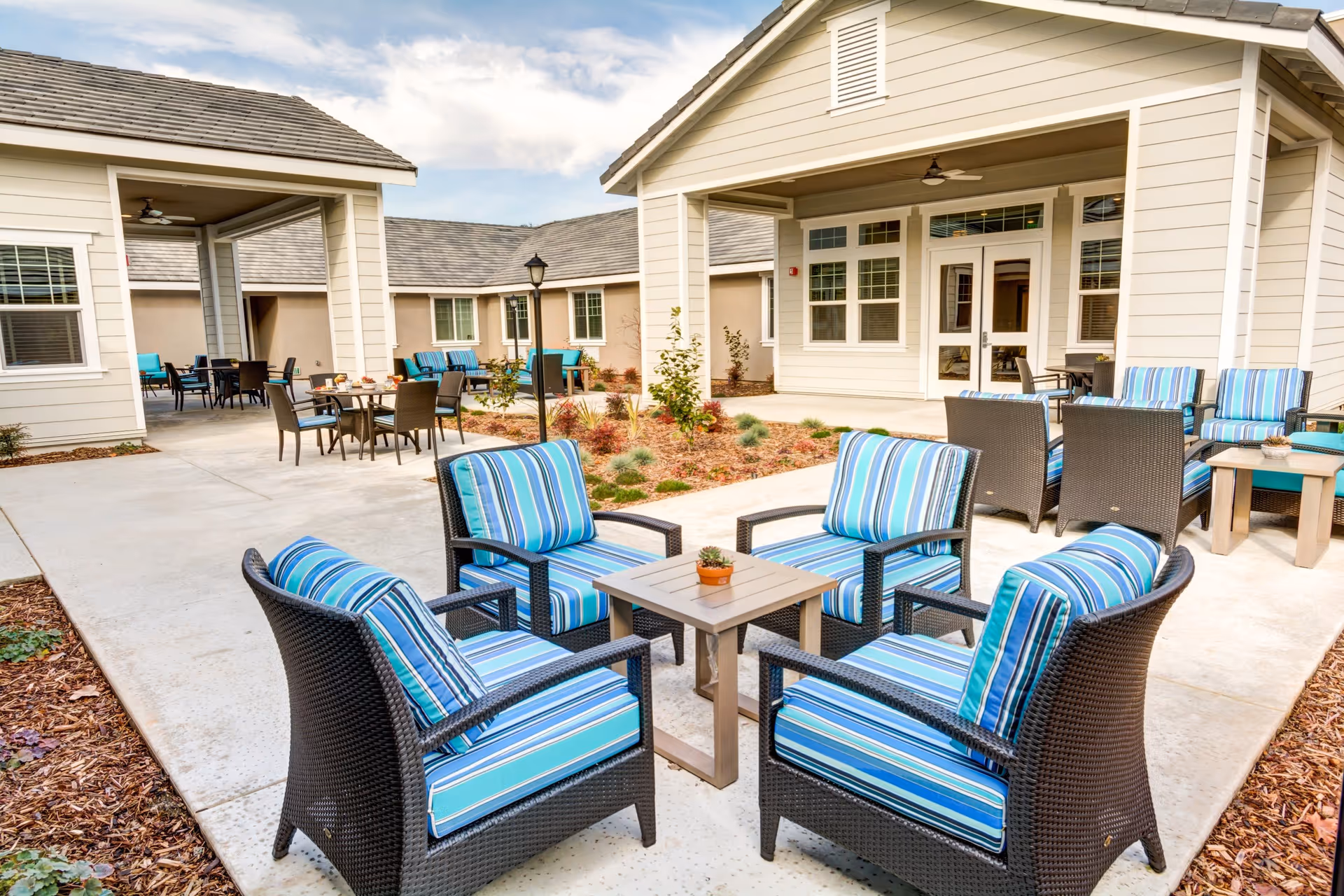 Outdoor patio area at The Courte At Citrus Heights featuring multiple seating arrangements with cushioned wicker chairs in blue striped fabric and small tables, surrounded by beige buildings and a partly cloudy sky.