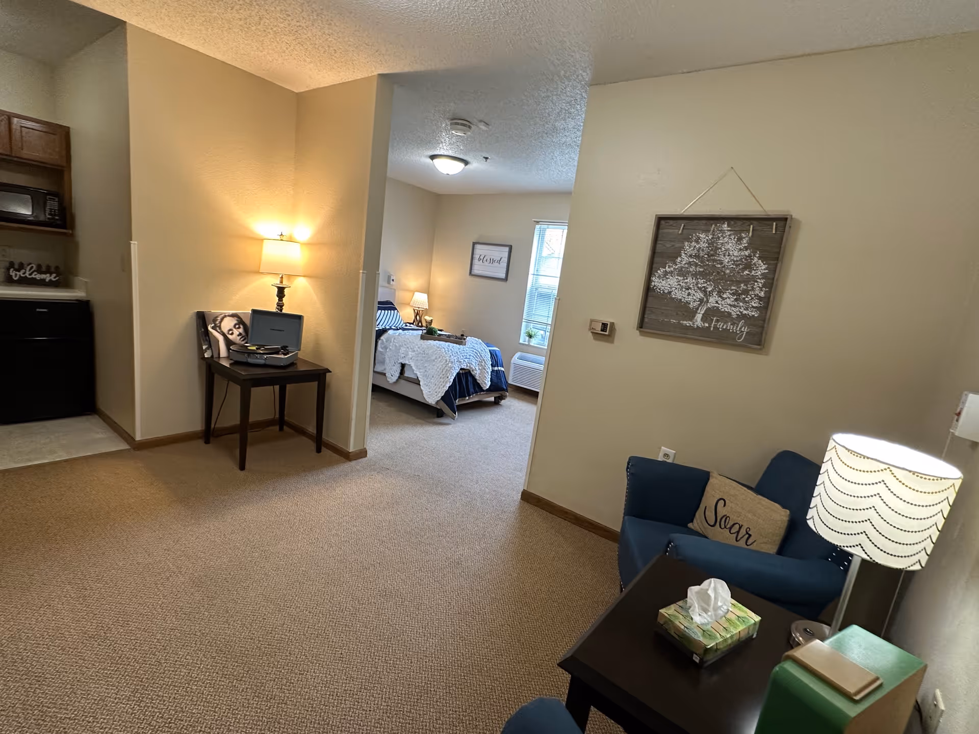 Interior view of a senior living facility apartment showing a small living area with a blue armchair, a side table with a lamp and tissue box, a wall decoration with a tree and the word 'Family', and a bedroom in the background with a bed, bedside table, and window. A kitchenette area is partially visible on the left with a microwave and cabinets.