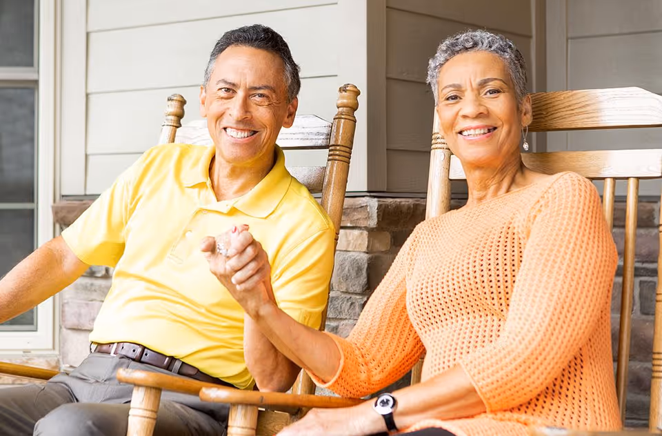 An elderly man and woman sitting on wooden rocking chairs on a porch, smiling and holding hands. The man is wearing a yellow polo shirt and gray pants, and the woman is wearing an orange knit sweater and a watch. The background shows a stone wall and siding of the building.