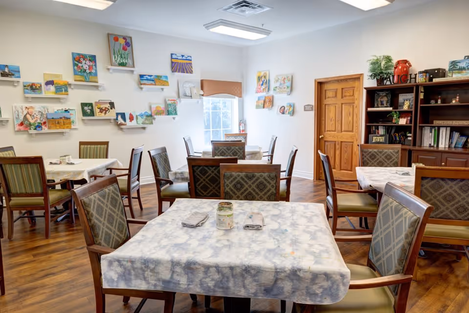 A bright room with several tables covered in white and blue patterned tablecloths, each surrounded by wooden chairs with patterned cushions. The walls are decorated with numerous colorful paintings displayed on small white shelves. There is a wooden door and a bookshelf filled with books and decorative items, including a sign that says 'LAUGH'. The floor is wooden, and a window allows natural light into the room.