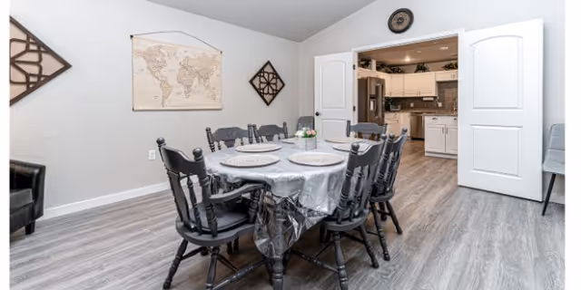A dining room with a table covered by a tablecloth surrounded by eight black chairs, wall decor, and double doors opening into a kitchen.