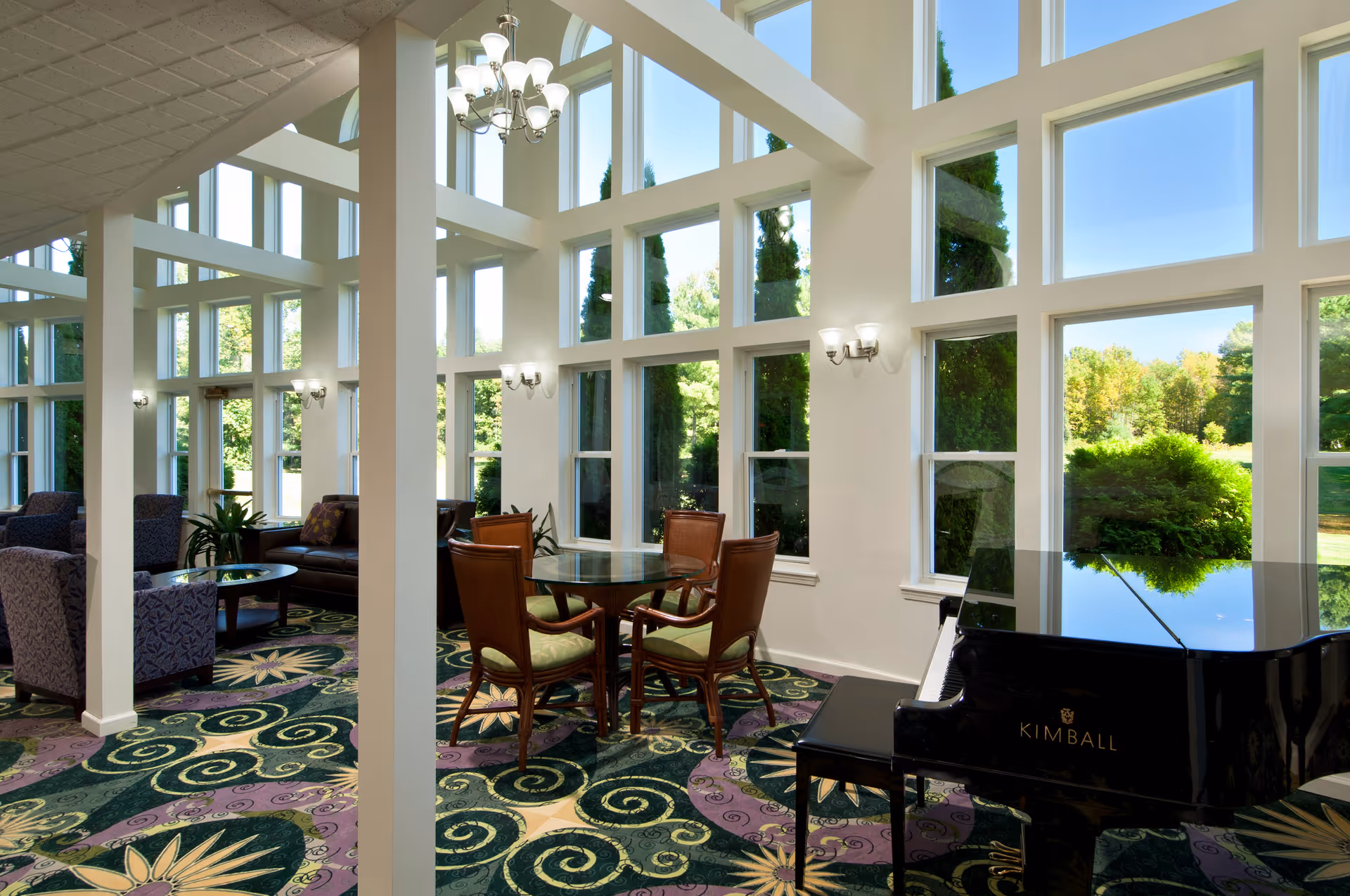 A bright and spacious living room area with large floor-to-ceiling windows letting in natural light. The room features a patterned carpet with green and purple swirls and floral designs, a black Kimball grand piano, a round glass table with four wooden chairs, and several upholstered armchairs and a sofa. The walls are white, and there are multiple wall sconces and a chandelier providing additional lighting. Outside the windows, green trees and a clear blue sky are visible.