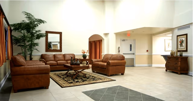 A spacious living room area with three brown leather sofas arranged around a wooden coffee table on a patterned rug. There is a large green plant in the corner, a mirror on the wall, and framed artwork above a wooden chest of drawers. The room has tiled floors and light-colored walls with a high ceiling.