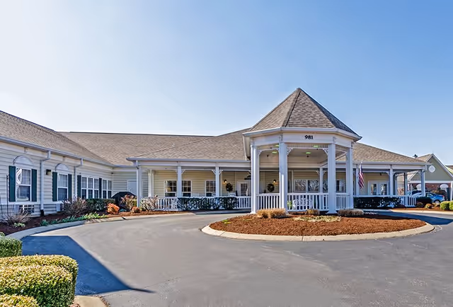 Front exterior view of The Bungalows at Bowling Green facility with a circular driveway, white railings, and a peaked roof entrance under a clear blue sky.