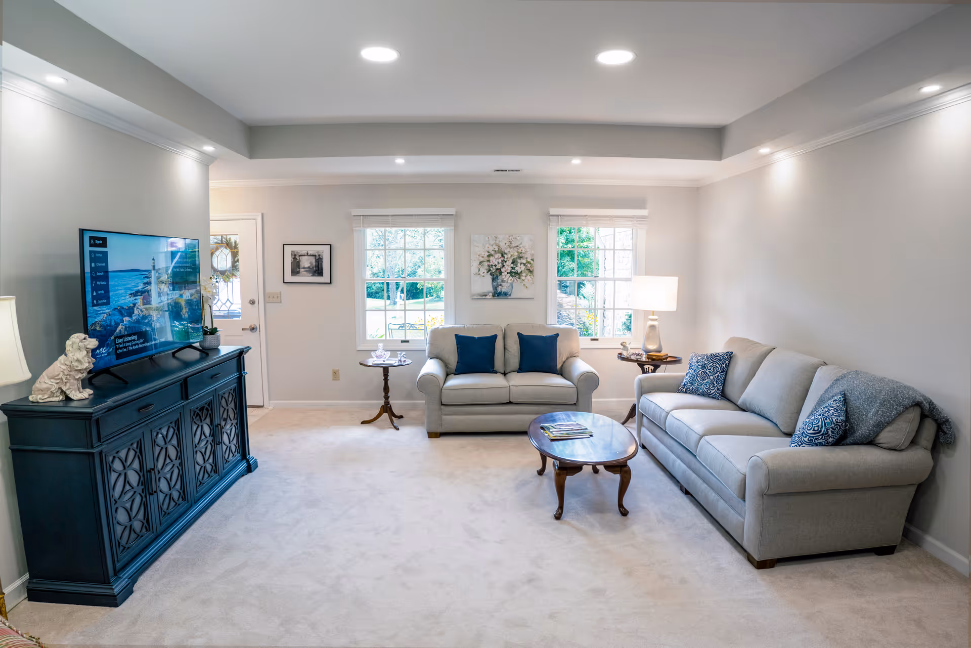 A bright and cozy living room with two light gray sofas adorned with blue and patterned throw pillows. A wooden coffee table sits in the center on a light carpeted floor. There is a dark blue TV stand with a flat-screen TV on top, and a small decorative dog statue. Two windows with white blinds let in natural light, and a table lamp on a side table adds warmth to the room. The walls are painted light gray with white trim, and there is a framed picture and a floral painting on the walls.