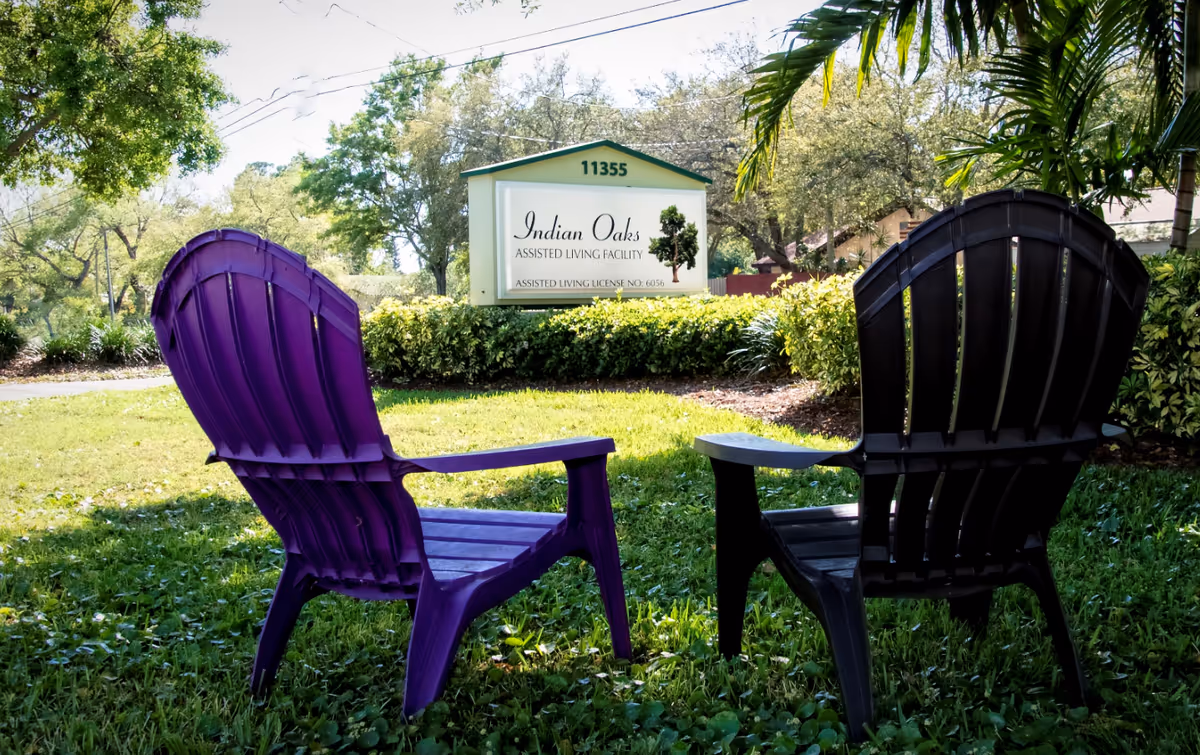 Two outdoor Adirondack chairs on a grassy lawn facing a sign for the Indian Oaks assisted living facility.
