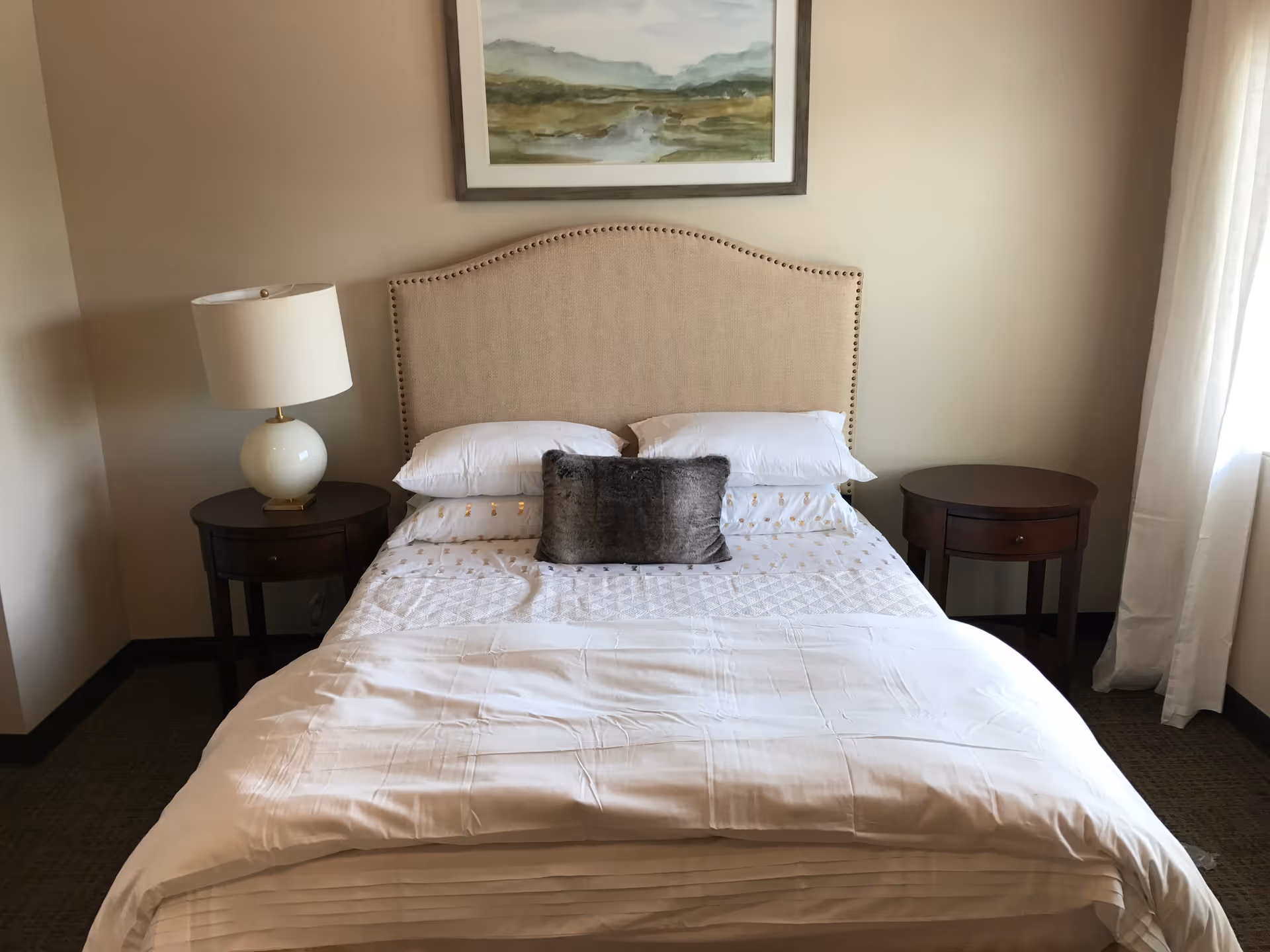 A neatly made bed with a beige upholstered headboard, white bedding, two white pillows, and a small gray decorative pillow in the center. There are two dark wooden nightstands on either side of the bed, with a white lamp on the left nightstand. A framed landscape painting hangs on the wall above the bed. Light beige walls and a window with white curtains are visible on the right side.