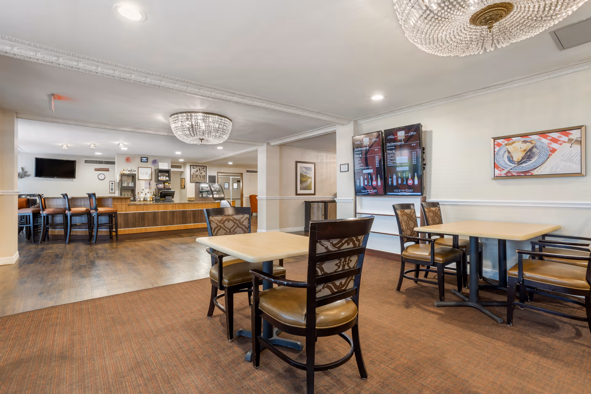 Interior view of a dining area in a senior living facility with wooden tables and chairs, a bar counter with stools, two digital menu boards on the wall, a framed picture of a slice of pie, and a decorative chandelier on the ceiling.