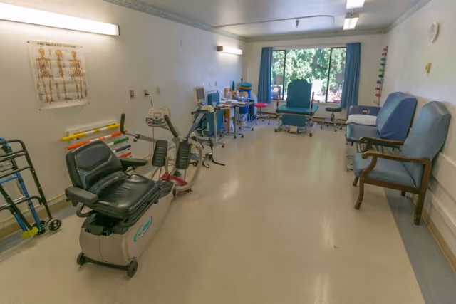 A rehabilitation therapy room with exercise equipment including a recumbent bike, parallel bars, and therapy chairs. The room has a large window with blue curtains letting in natural light, medical posters on the wall, and various therapy tools and chairs arranged along the walls.