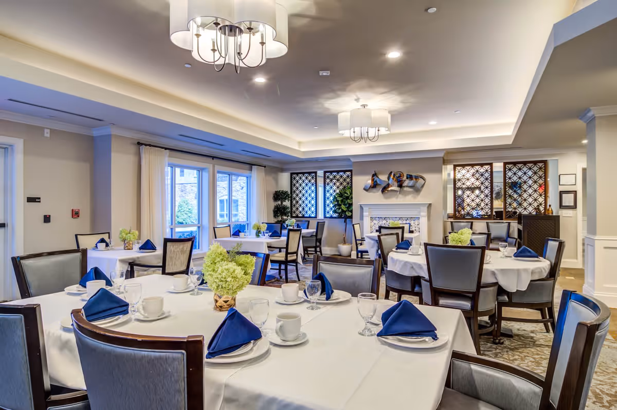A well-lit dining room with multiple round tables covered in white tablecloths, each set with white cups, plates, glasses, and blue folded napkins. The room features large windows with white curtains, decorative plants, modern chandeliers, and a fireplace with artistic wall decor above it.