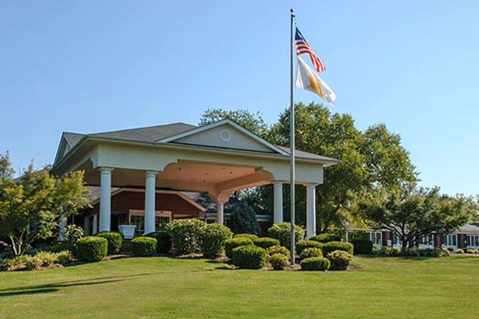 Exterior view of a senior living facility building with a covered entrance supported by white columns, surrounded by well-maintained bushes and trees, with an American flag and another flag flying on a flagpole in front of the building under a clear blue sky.