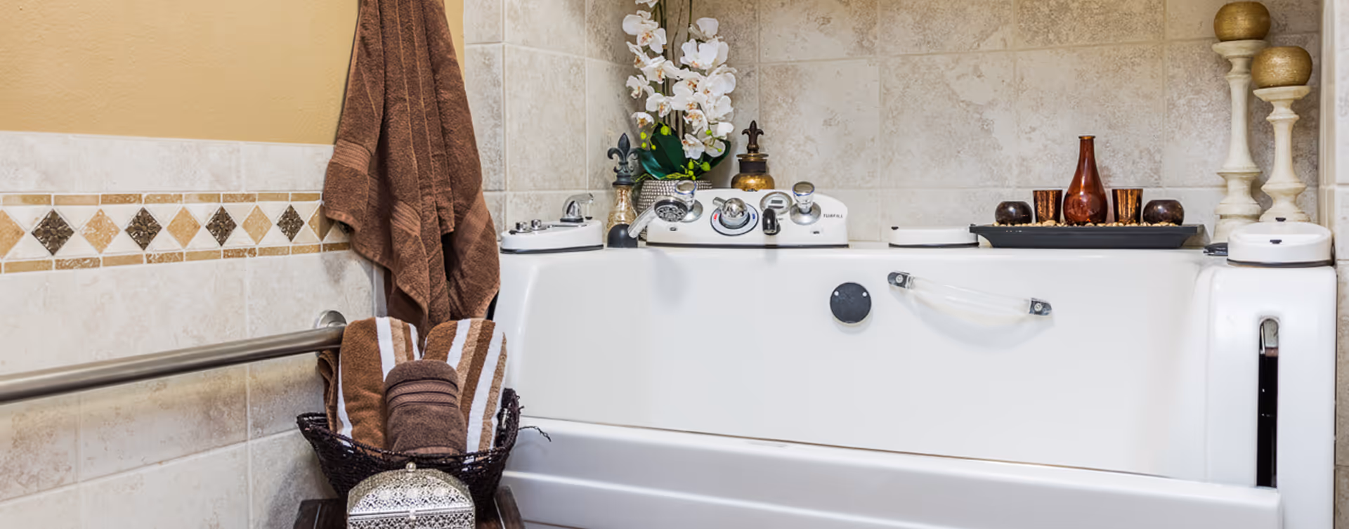 A bathroom featuring a white walk-in bathtub with chrome fixtures and a safety grab bar. The tub is surrounded by beige tiled walls with decorative diamond-shaped accents. Brown towels hang on a rail and are neatly rolled in a basket beside the tub. Decorative items including white orchids, candles, and vases are placed on the tub ledge.