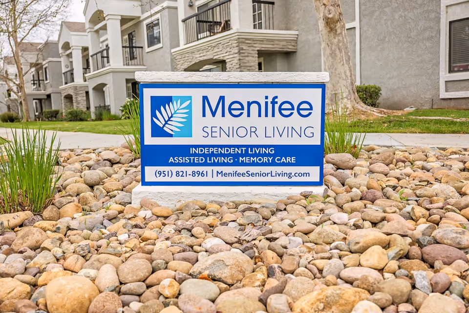 A blue and white "Menifee Senior Living" sign sits on a bed of rocks in front of an apartment building.