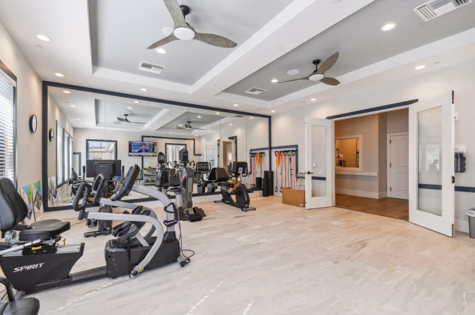 Bright fitness room with exercise bikes, large wall mirror, ceiling fans, and resistance bands hanging on the wall. The room has light-colored flooring and white walls with recessed lighting. Double glass doors open to an adjacent hallway.